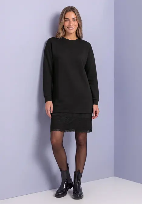 Frau in schwarzem Strickkleid mit Spitzenbesatz, schwarzen Strumpfhosen und Stiefeletten vor hellvioletter Wand.
