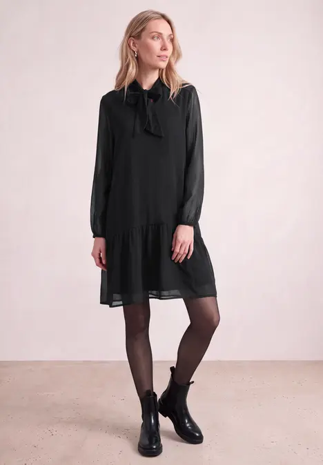 Eine Frau vor hellem Hintergrund in schwarzem Kurzkleid mit Schleife, schwarzen Strumpfhosen und Stiefeletten.