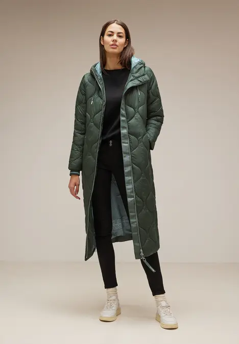Frau mit langem, grünem Steppmantel, schwarzer Hose und weißen Sneakers.