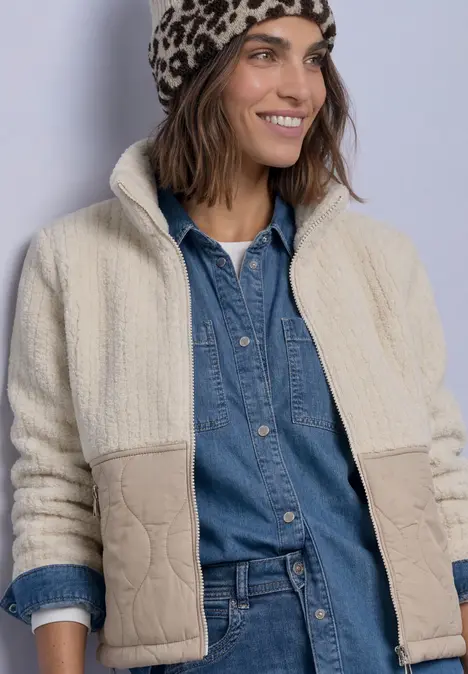 Frau in hellem Teddy-Fleece-Jacke, Jeanshemd und Leopardenmütze lächelt.