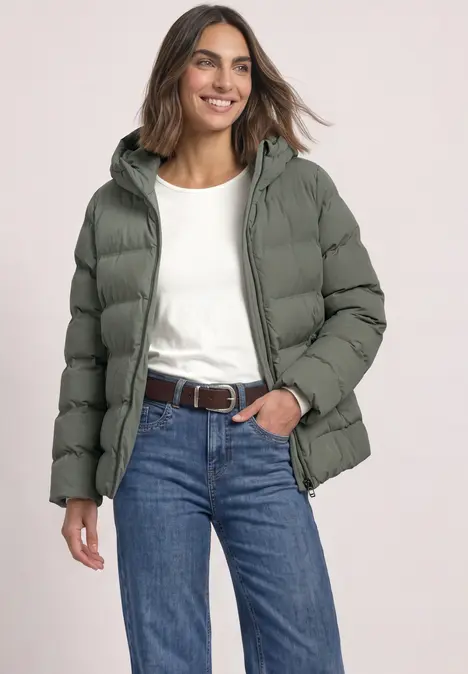 Femme portant une doudoune gris-vert à capuche, un t-shirt blanc, un jean et une ceinture, souriante