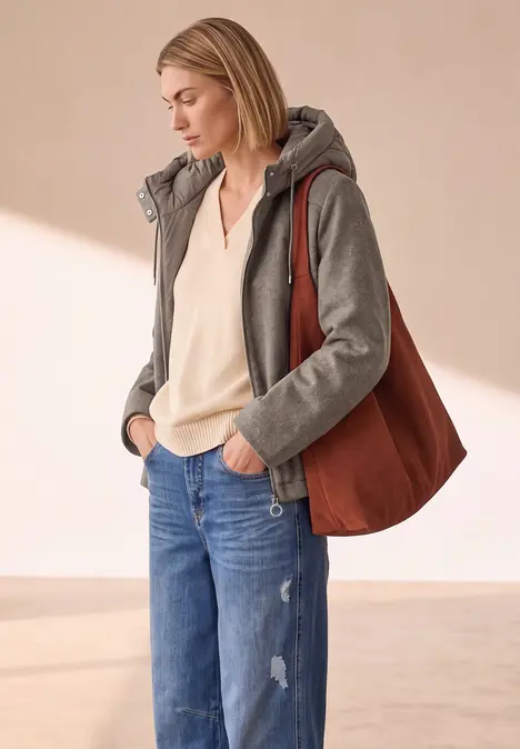 Frau in grauer Kapuzenjacke, weißer Bluse, blauen Jeans und brauner Schultertasche, stehend vor hellem Hintergrund.