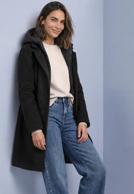 Vrouw in een zwarte jas, licht truitje en blauwe jeans, glimlachend voor een neutrale achtergrond.