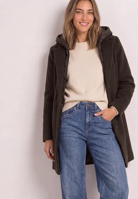 Junge Frau trägt braunen Parka über hellem Pullover und blauen Jeans, posiert lässig.