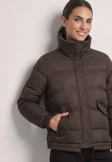 Frau trägt eine braune gesteppte Jacke mit hohem Kragen und steckt die Hände in die Taschen.