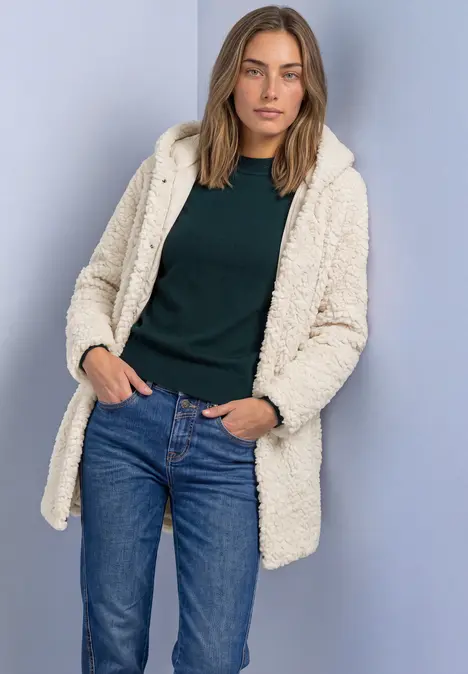 Kobieta w kremowym płaszczu teddy, ciemnozielonym topie i niebieskich jeansach na neutralnym tle.