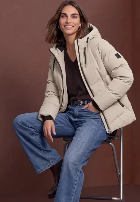 Frau trägt beige Steppjacke, schwarzen Pullover und Jeans, sitzt auf Hocker.