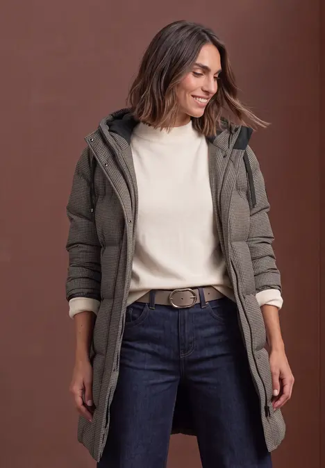 Frau mit khakigrünem Winterparka, cremefarbenem Pullover und Jeans, lächelnd seitlich vor braunem Hintergrund.