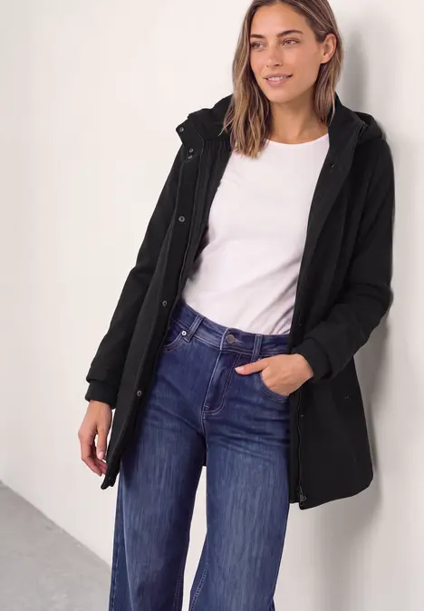 Frau trägt schwarzen Mantel, weißes T-Shirt und Jeans, steht vor heller Wand, eine Hand in Tasche, andere hält Handy.