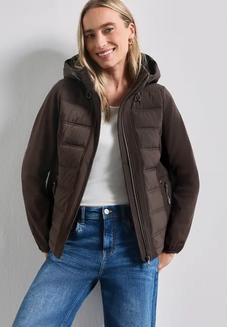 Frau in weißem Top, blauer Jeans und brauner gesteppter Jacke, lächelnd vor neutralem Hintergrund.