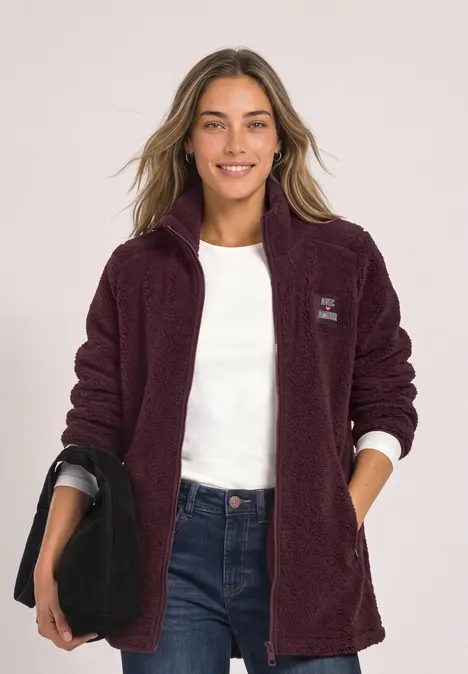 Femme en veste polaire bordeaux, chemise blanche, jean, tenant un sac noir.