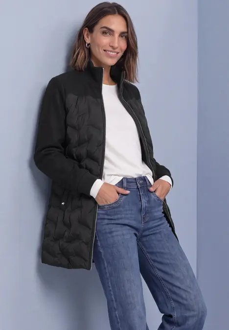 Femme en doudoune noire, top blanc et jean bleu, souriante, mains dans les poches sur fond gris.