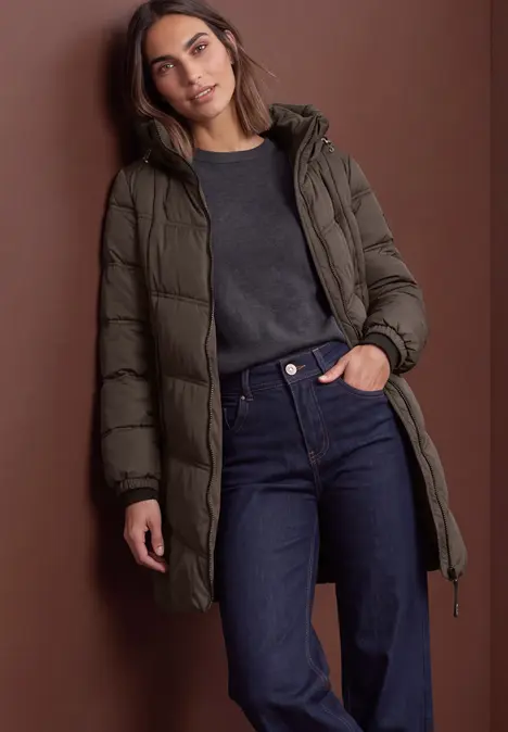 Frau in olivgrünem Daunenmantel, dunklem Pullover und Jeans vor braunem Hintergrund.