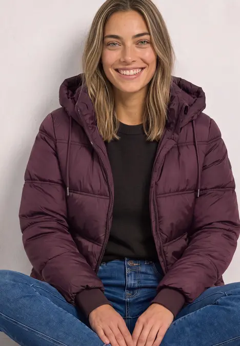 Eine lächelnde Frau sitzt mit verschränkten Beinen und trägt eine violette Steppjacke, dunklen Pullover und Jeans.