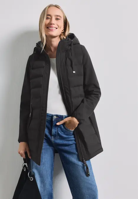 Frau mit schwarzer Winterjacke, Kapuze, Jeans, lächelnd vor weißem Hintergrund.