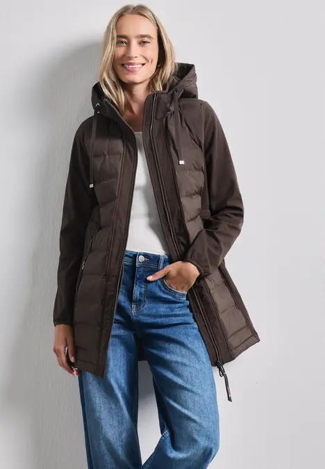 Eine Frau trägt eine braune Steppjacke mit Kapuze, weiße Bluse und blaue Jeans.