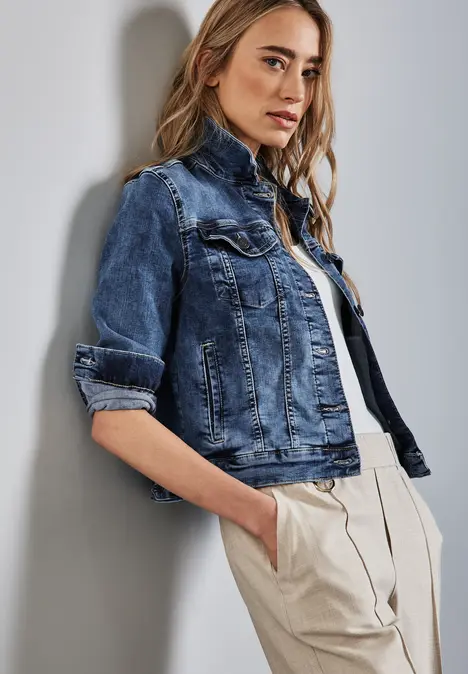 Frau mit blauer Jeansjacke und beiger Hose lehnt an einer Wand.