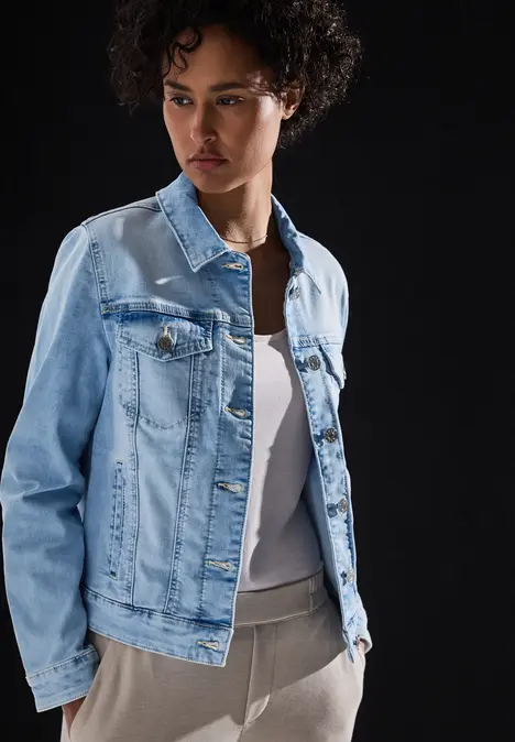 Frau mit lockigem Haar trägt helle Jeansjacke und weißes Top vor schwarzem Hintergrund.