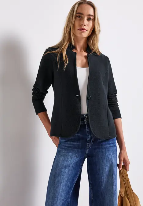 Frau in schwarzem Blazer, weißem Shirt und blauer Jeans steht vor weißer Wand.