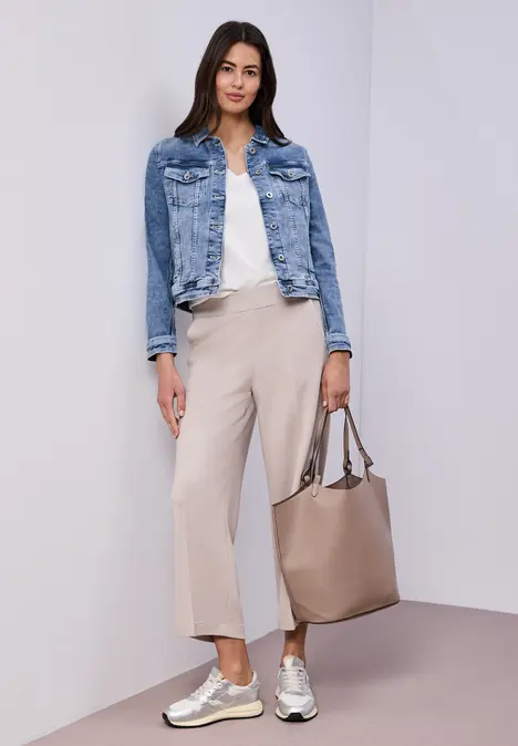 Frau in Jeansjacke, weißem Top, hellbeigen Culottes und Sneakers, mit taupefarbener Tasche vor neutralem Hintergrund.
