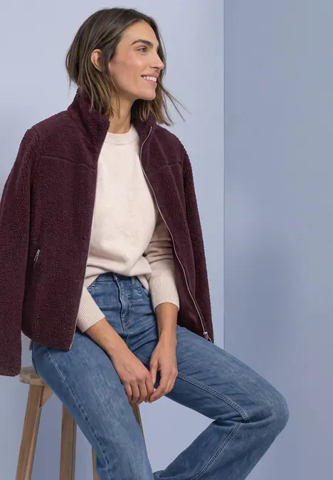 Frau sitzt lächelnd auf Hocker, trägt bordeauxfarbenes Fleece-Jacke, hellen Pullover und Jeans.