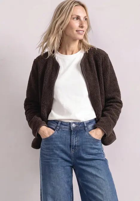 Frau mit brauner Teddyjacke, weißem T-Shirt und blauen Jeans, Hände in den Taschen, seitlicher Blick.