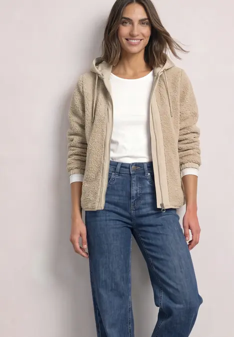 Eine Frau trägt eine beige Teddyjacke, weißes Shirt und blaue Jeans und lächelt entspannt.