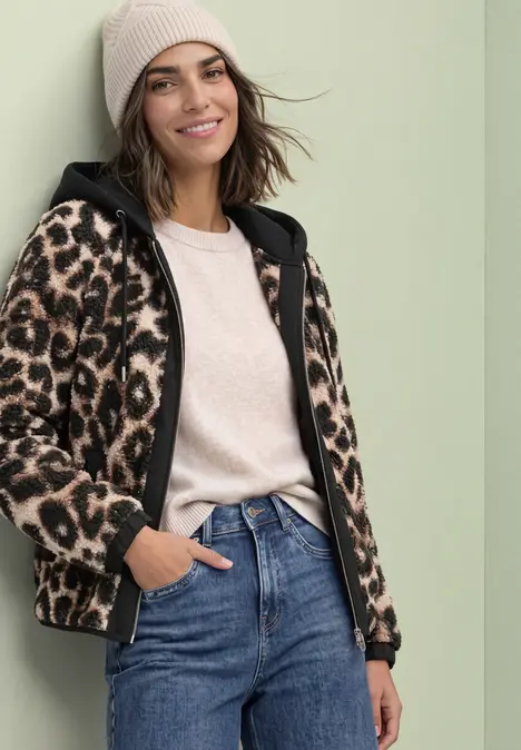 Vrouw in jeans, beige muts en luipaard-fleecejas lacht ontspannen.