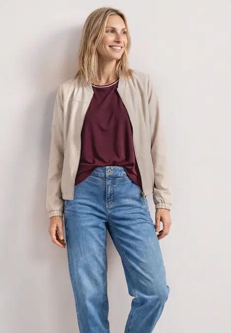 Eine Frau trägt eine beige Jacke, burgunderfarbenes Shirt und blaue Jeans vor weißer Wand.