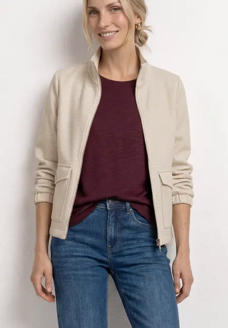 Frau steht vor weißem Hintergrund, trägt beige Bomberjacke, bordeauxrotes T-Shirt und blaue Jeans.