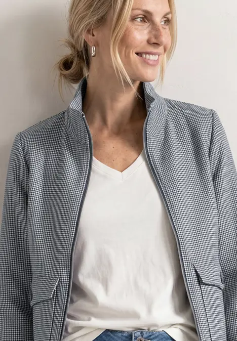 Frau in hellgrauer Jacke mit Reißverschluss, weißem T-Shirt und Ohrsteckern, Haare locker hochgesteckt.