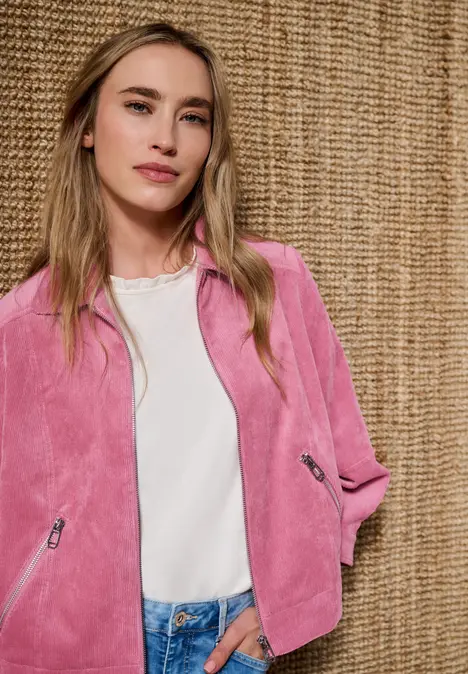 Eine Frau in rosa Jacke über weißem Shirt und Jeans steht vor einem gewebten Hintergrund.