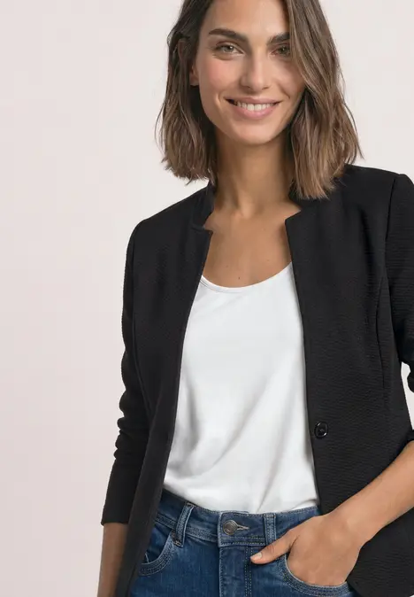 Eine lächelnde Frau in schwarzem Blazer und weißem Shirt, Hand in der Hosentasche.