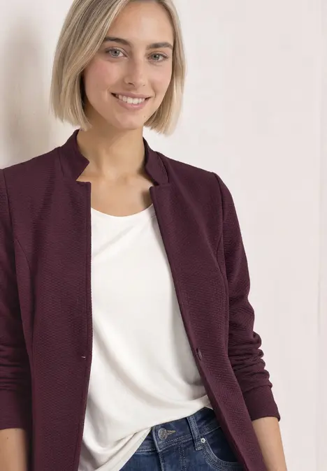 Eine lächelnde Frau mit kurzem blonden Haar, trägt eine dunkelrote Strickjacke, weißes Shirt und Jeans.
