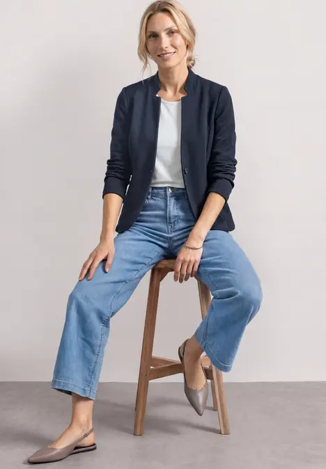 Frau sitzt auf Hocker, trägt weiße Bluse, dunkelblaue Jacke, weite Jeans und graue Ballerinas.