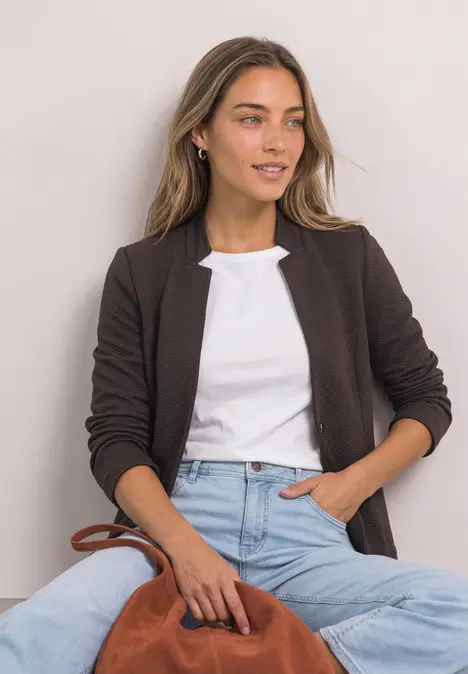 Junge Frau trägt schwarzen Blazer, weißes Shirt und Jeans, hält eine braune Tasche.