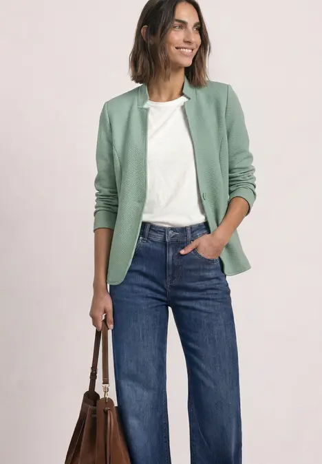 Frau in grünem Blazer, weißem Shirt, weiten Jeans mit Handtasche, lächelnd vor neutralem Hintergrund.