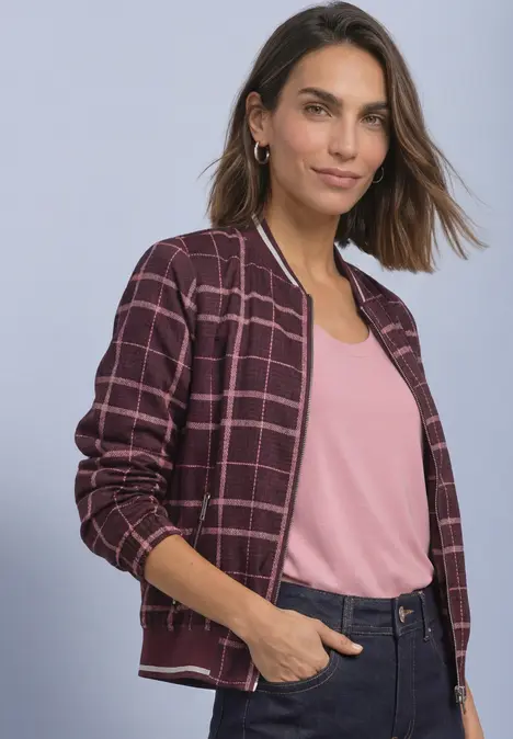 Frau trägt karierte, bordeauxfarbene Bomberjacke zu rosa Top und dunkler Jeans vor neutralem Hintergrund.