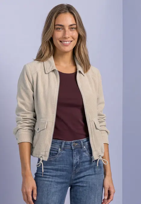 Eine lächelnde Frau in beigefarbener Jacke, bordeauxrotem Shirt und Jeans vor hellviolettem Hintergrund.