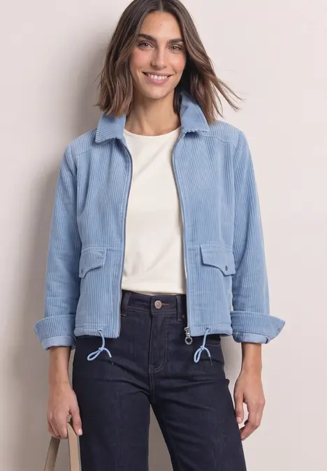 Frau in blauer Jacke, weißem Shirt und dunklen Jeans, lächelnd.