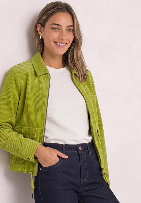 Junge Frau in grüner Jacke, weißem Shirt und Jeans, die lächelnd in die Kamera blickt.