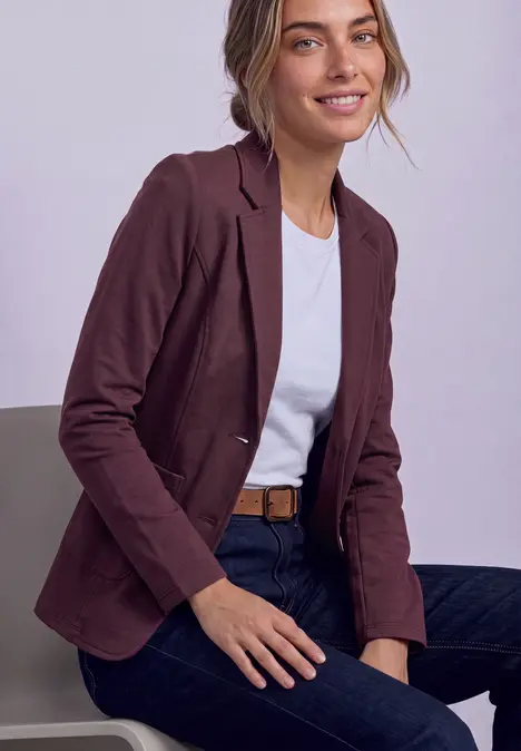 Frau in weißem T-Shirt, dunkelrotem Blazer, Jeans und Gürtel sitzt lächelnd.