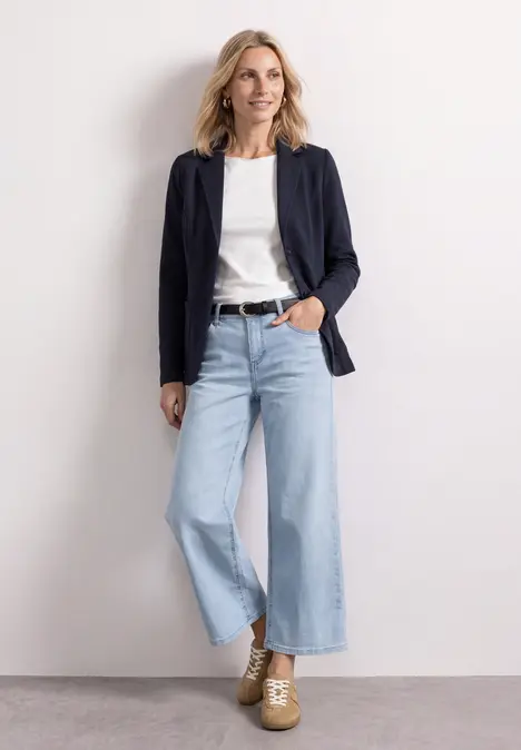 Frau im casual Chic: weiße Bluse, navy Blazer, weite Jeans und Plateau-Schuhe, lächelnd vor hellem Hintergrund.