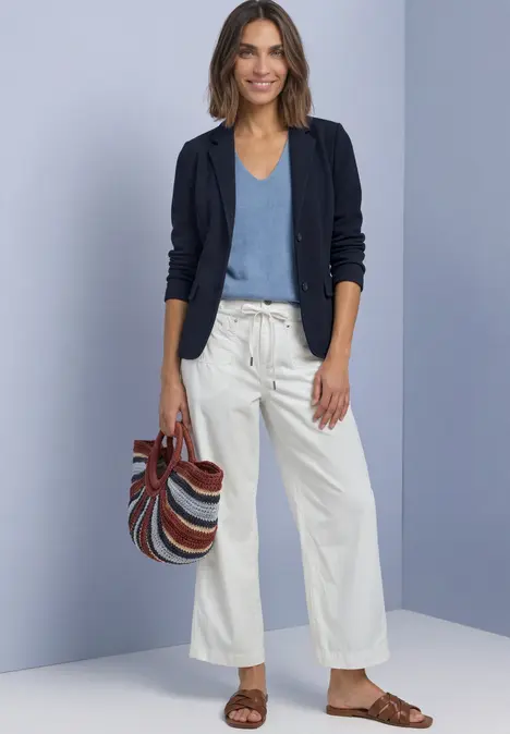 Frau mit dunkelblauem Blazer, hellblauem Top, weißer Culotte, braunen Sandalen und gestreifter Tasche.