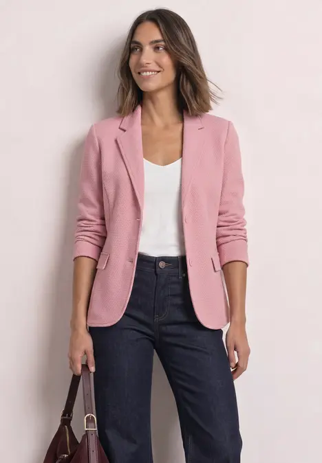 Frau im rosa Blazer, weißem Shirt und dunklen Jeans, hält eine Handtasche vor hellem Hintergrund.