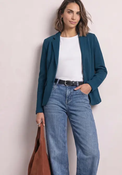 Vrouw met een teal blazer, wit shirt, jeans en een bruine tas