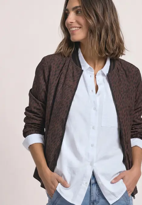 Frau lächelt in einer locker sitzenden Strickjacke mit abstraktem Muster über weißer Bluse, Hände in Taschen.