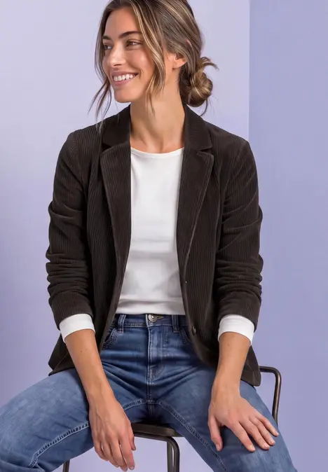 Vrouw zit glimlachend op een stoel, draagt een zwarte blazer, een wit shirt en een jeans tegen een lichte achtergrond.