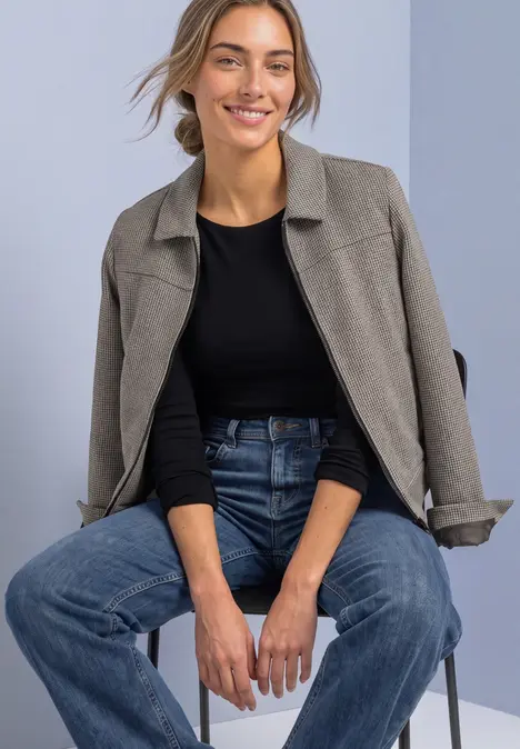Lächelnde Frau in Jeans, schwarzem Shirt und grauer Jacke sitzt auf einem Stuhl in schlichtem Studio-Setting.