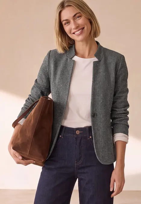 Lächelnde Frau in grauem Blazer, weißem T-Shirt, dunkelblauer Jeans und brauner Umhängetasche.